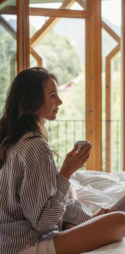 Frau sitzt entspannt mit Kaffee auf dem Bett und blickt aus dem Fenster in die Natur.