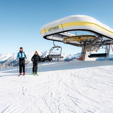 Zwei Skifahrer vor Sesselliftstation in verschneiter Berglandschaft bei strahlend blauem Himmel.